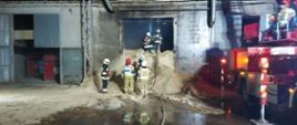 Zdjęcie przedstawia pomieszczenie, w którym doszło do pożaru. Widoczne są składowane trociny, strażacy, którzy wykonują swoje czynności gaśnicze oraz po prawej stronie widoczny jest podnośnik hydrauliczny.