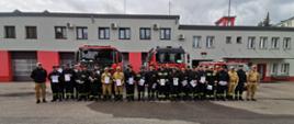 Wspólne zdjęcie strażaków OSP po zakończonym egzaminie, na tle wozów strażackich przed budynkiem komendy.