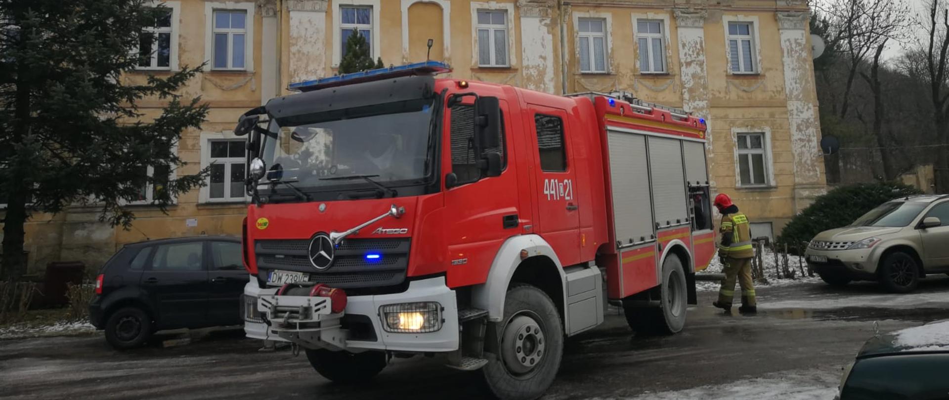 Obraz przedstawia pojazd pożarniczy z włączonymi światłami alarmowymi. Pojazd zaparkowany na ulicy. W tle budynek mieszkalny wielorodzinny.