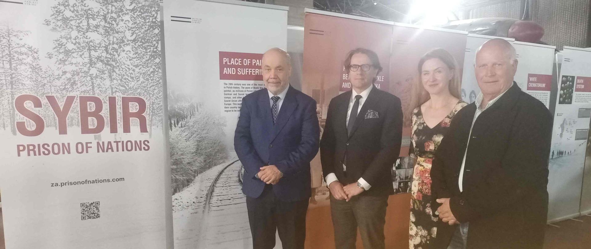 A group of four people pose for a photo; from left, Chargé d'Affaires Jacek Chodorowicz, Director of the Sybir Memorial Museum, Prof. Wojciech Śleszyński, Julita Waś, and President of the Polish Association of Siberian Deportees of South Africa, Stefan Szewczuk; posters from the "Sybir: Prison of Nations" exhibition in the background.