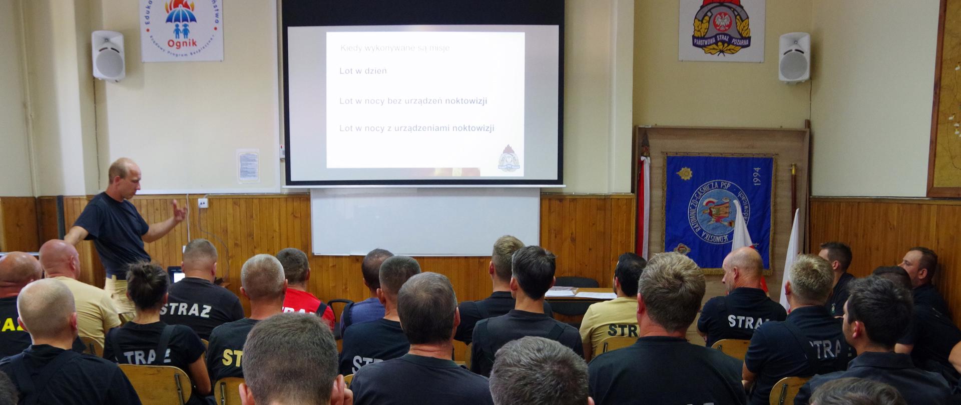 Widok z tyłu. Na pierwszym planie strażacy OSP siedzą na sali szkoleniowej. W tle po lewej stronie instruktor z PSP a na środku na ekranie obraz z rzutnika.