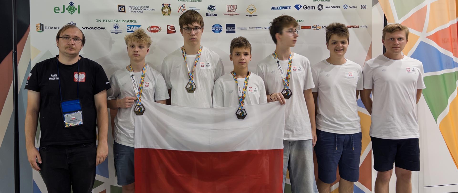 
Grupa sześciu młodych mężczyzn i jednego dorosłego mężczyzny trzymających polską flagę. Młodzi mężczyźni mają na szyjach zawieszone medale. W tle widoczny jest baner z napisem „EJOI 2025 European Junior Olympiad in Informatics Shumen, Bulgaria 29 August – 04 September”.