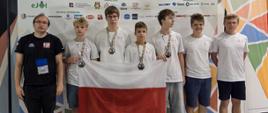 Grupa sześciu młodych mężczyzn i jednego dorosłego mężczyzny trzymających polską flagę. Młodzi mężczyźni mają na szyjach zawieszone medale. W tle widoczny jest baner z napisem „EJOI 2025 European Junior Olympiad in Informatics Shumen, Bulgaria 29 August – 04 September”.