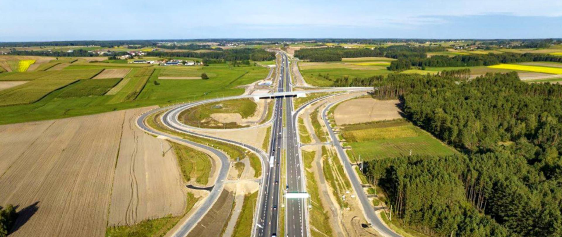 Widok z lotu ptaka na węzeł drogowy na trasie S61, z dwujezdniową drogą ekspresową i zakrętami łącznic, otoczony polami i terenami zielonymi.