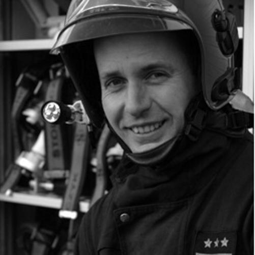 uśmiechnięty strażak ubrany w mundur specjalny i hełm strażacki, w tle sprzęt pożarniczy
