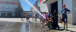 Zdjęcie przedstawia czternaścioro dzieci oraz dwóch strażaków podczas polewania wodą z węża strażackiego na placu wewnętrznym komendy
