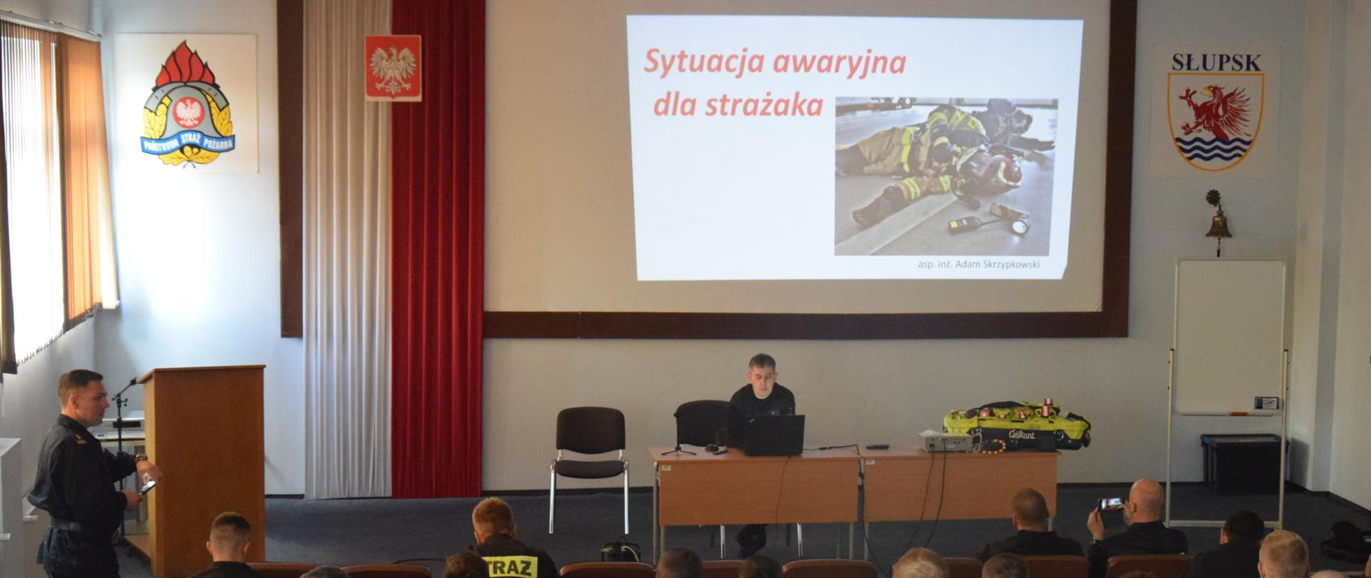 Strażacy siedzą na fotelach podczas prowadzenia zajęć przez dwóch funkcjonariuszy Państwowej Straży Pożarnej.