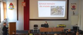 Strażacy siedzą na fotelach podczas prowadzenia zajęć przez dwóch funkcjonariuszy Państwowej Straży Pożarnej.