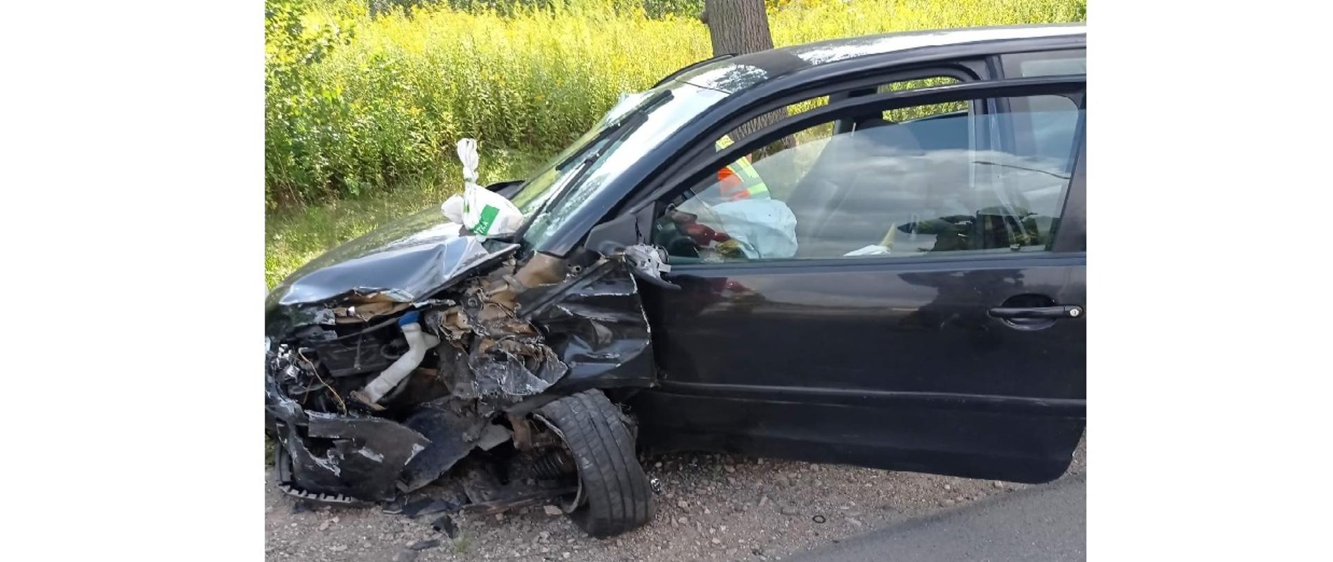 Na środku zdjęcia widać uszkodzony czarny samochód osobowy, którego lewy błotnik oraz koło w wyniku zdarzenia uległo całkowitemu zniszczeniu. Uszkodzony również został przedni zderzak oraz maska. Pojazd ma otwarte drzwi od strony kierowcy. W środku pojazdu widać wystrzeloną kurtynę powietrzną z kierownicy pojazdu. Na masce przy szybie leży biało zielona foliowa reklamówka. Przed pojazdem widoczna jest część drogi asfaltowej. Za pojazdem w centralnej części zdjęcia widać drzewo, a w oddali porasta zielona roślinność oraz mniejsze drzewka liściaste.