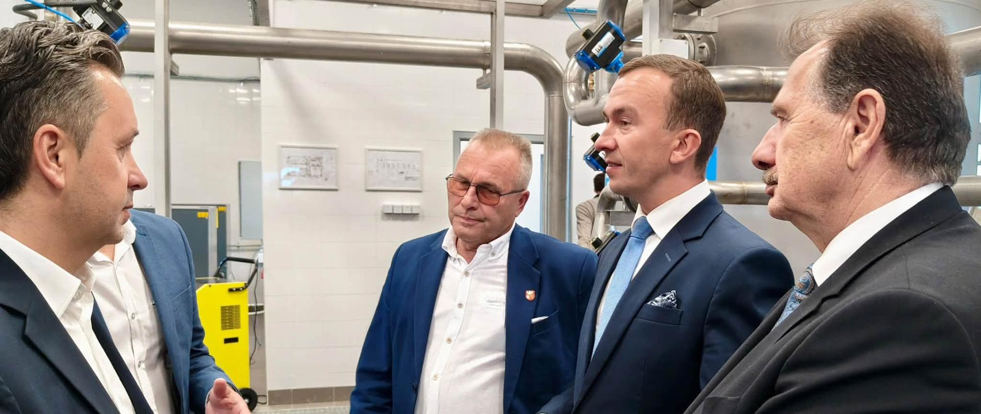 Czterech mężczyzn w garniturach rozmawia w fabryce, otoczeni maszynami i sprzętem przemysłowym. 