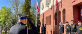 Strażak w mundurze galowym oddaje honor przed nim stoją strażacy w mundurze galowym oraz koszarowym strażacy obok masztu wciągają flagę Polski za nimi jest budynek komendy.