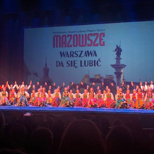 Zdjęcie sceny finałowej występu Państwowego Zespołu Ludowego Pieśni i Tańca Mazowsze w Teatrze Wielkim Operze Narodowej w Warszawie. Tancerki klęczą na jednym kolanie, z tyłu tancerze z podniesionymi rękoma ku górze.