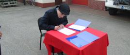 Przy stole pokrytym czerwonym suknem siedzi strażak ubrany w mundur służbowy. Na głowie ma założony beret. W ręku trzyma długopis, którym podpisuje akt ślubowania.