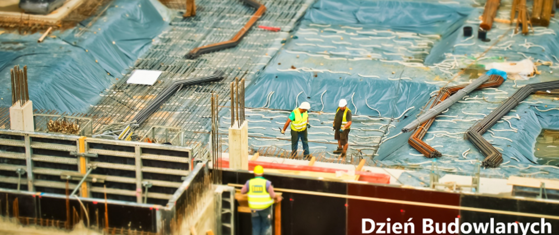 Robotnicy na placu budowy z niebieskimi plandekami i materiałami budowlanymi; na pierwszym planie bariera z napisem „Dzień Budowlanych”.