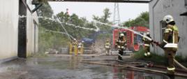 Teren zakładu. Na palący się budynek strażacy w pełnych ubraniach bojowych podają trzy prądy wody. W tle czerwony wóz bojowy.