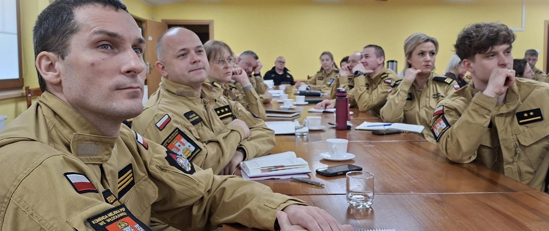 Przy stole obrad kilkunastu strażaków w mundurach służbowych, w kolorze piaskowym. Na stole dokumenty, filiżanki i szklanki. 