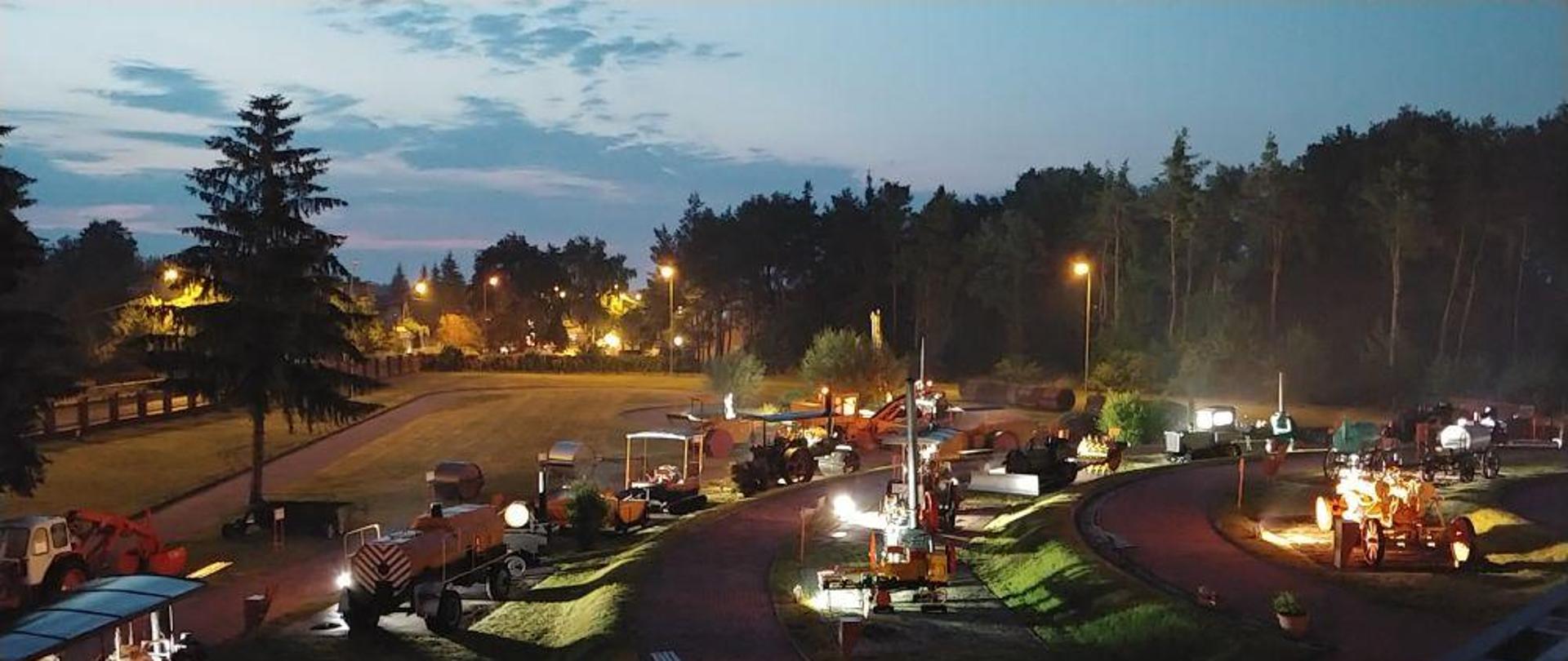 Nocne zdjęcie terenu wokół budynku muzeum. Widoczne eksponaty muzealne, drzewa i oświetlenie z lamp