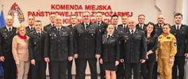 Strażacy w mundurach wyjściowych oraz pracownicy cywilni komendy stoją na tle białej ściany z czerwonym napisem Komenda Miejska Państwowej Straży Pożarnej w Skierniewicach