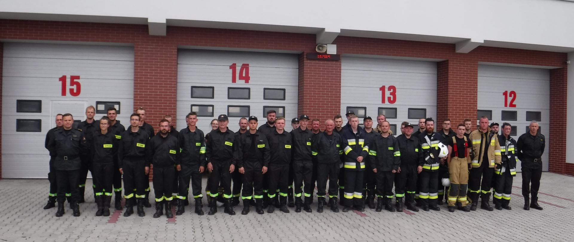 Kilkudziesięciu mężczyzn ubranych w czarne ubrania koszarowe strażackie z odblaskowymi paskami, czarnym butami, stoją na kostce betonowej przed bramami garażowymi oznaczonymi czerwonymi numerami. W tle budynek Komendy Powiatowej Państwowej Straży Pożarnej w Bochni. Budynek w kolorze białym, okna i drzwi koloru białego