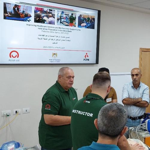 Grupa mężczyzn w pomieszczeniu stoi wokół szafki, na której stoi sprzęt elektroniczny. Na ścianie na monitorze widać zdjęcia logo Polskiej pomocy i PCPM. 