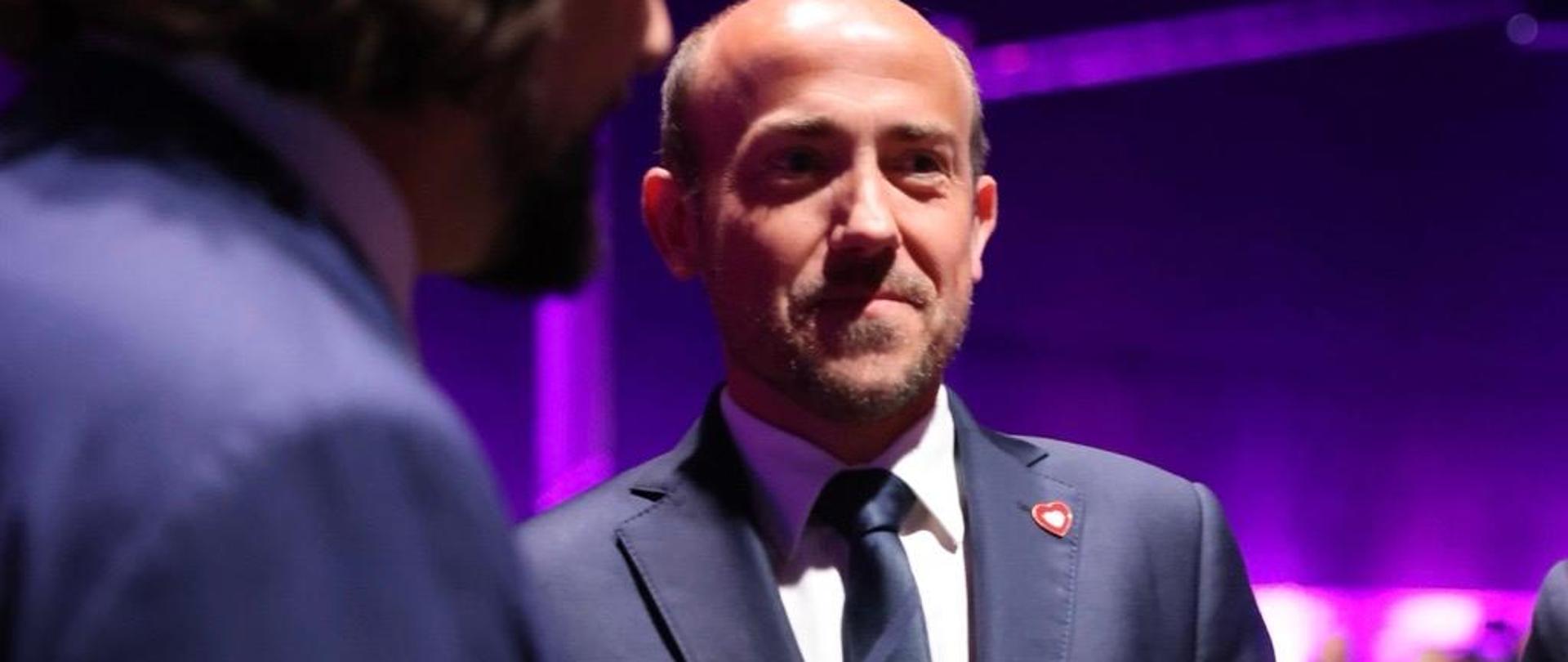 Zdjęcie przedstawia Ministra Borysa Budkę podczas Europejskiego Kongresu Gospodarczego. Na pierwszym planie, w nieostrości stoi mężczyzna, którego widoczny jest profil. Na drugim planie, w mocnym bocznym świetle Minister Borys Budka.