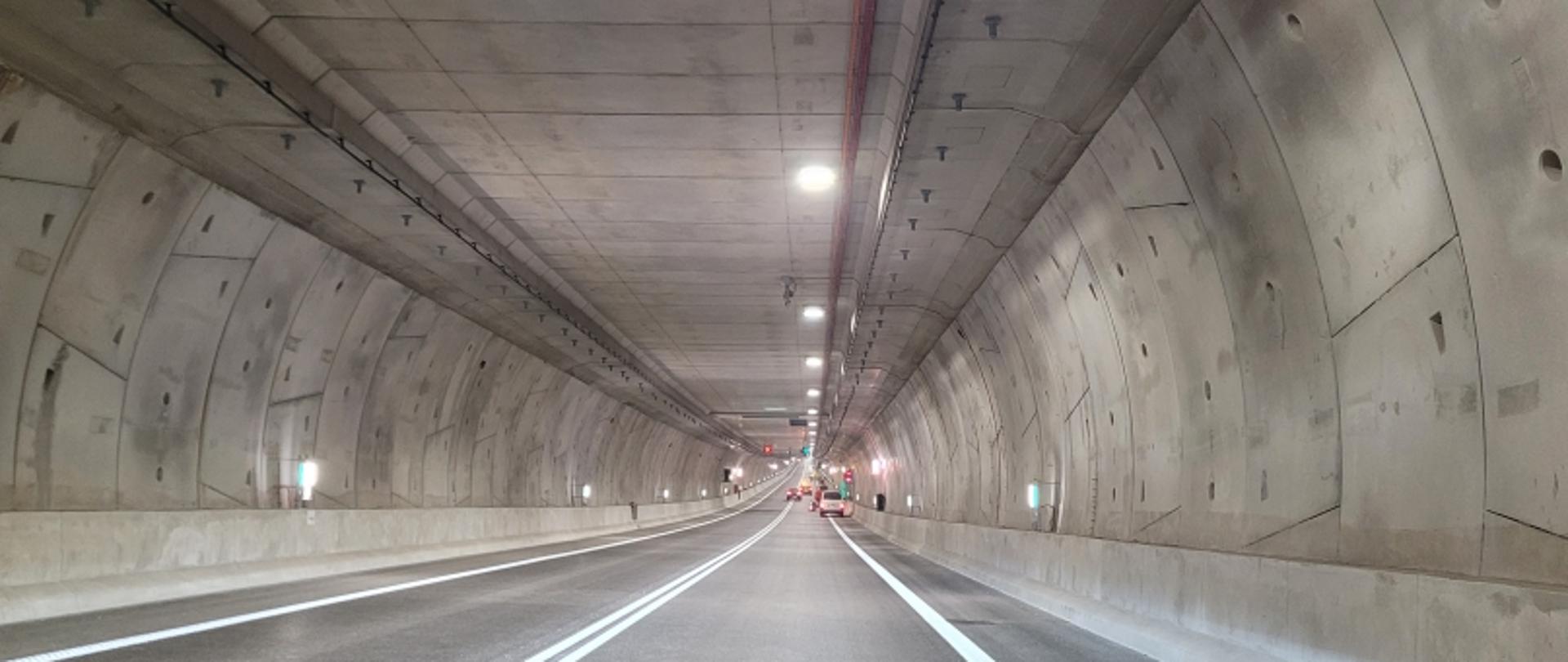 Tunel drogowy z dwoma pasami, betonowe ściany z panelami, oświetlenie sufitowe, kilka pojazdów w tle.