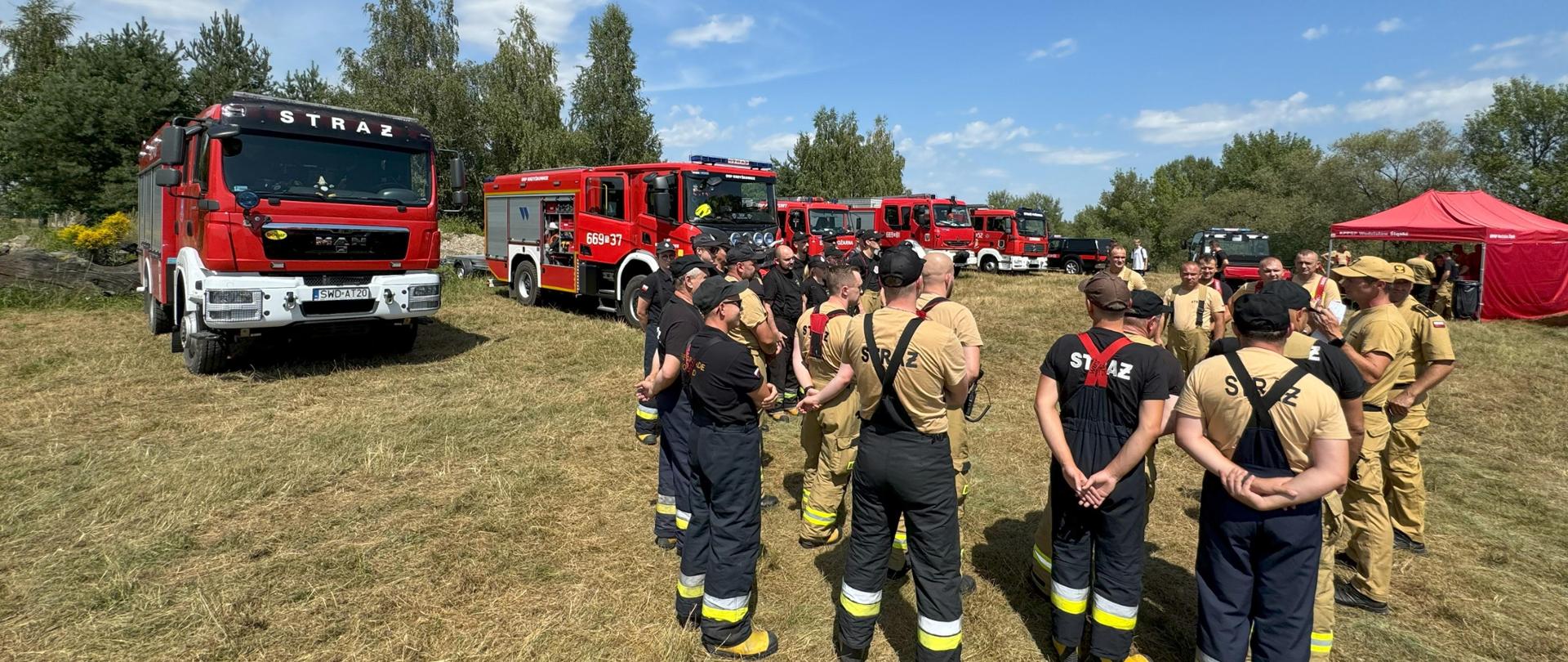 Na zdęciu zbiórka strażaków przed rozpoczęciem ćwiczeń. W tle samochody strażackie.