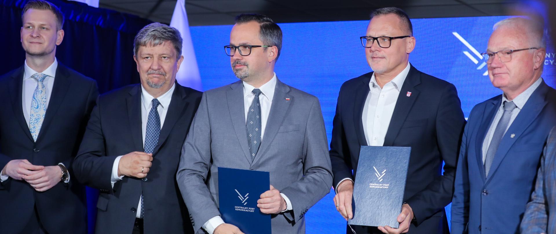 Pięć osób stoi obok siebie. W środku z teczką w rękach stoi wiceminister Marcin Horała. 