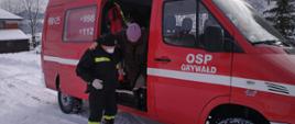 na zdjęciu na zaśnieżonej drodze strażak pomaga starszej pani wysiąść z pojazdu OSP Grywałd w celu udania się przez nią na szczepienie przeciwko COVID