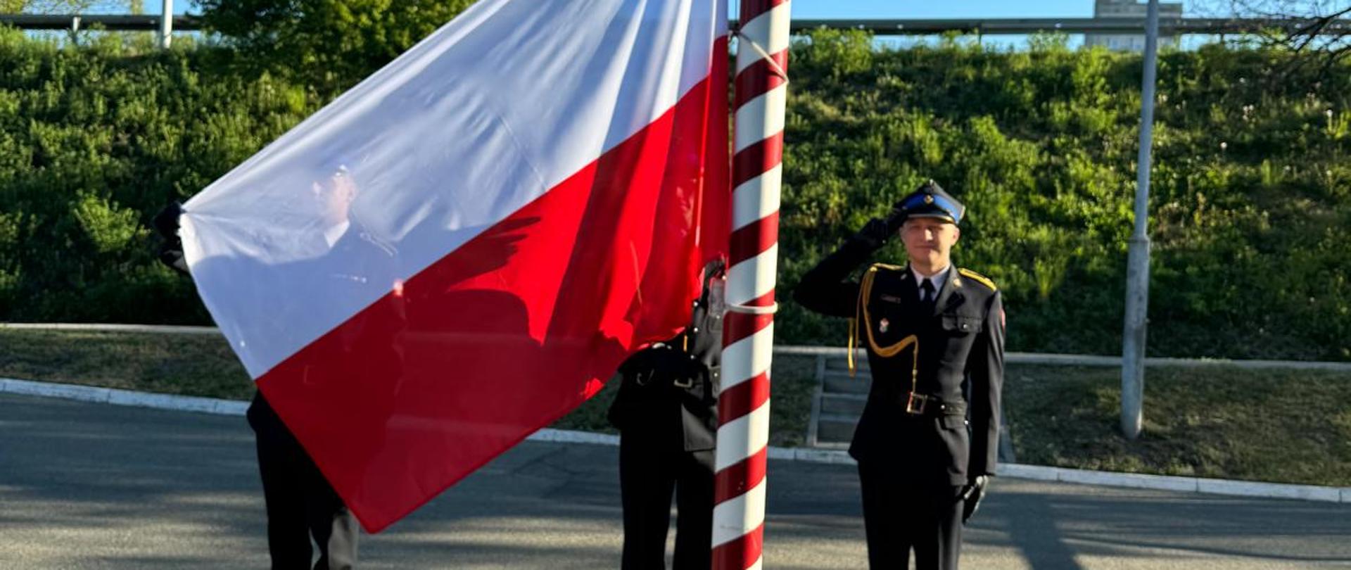 Zdjęcie przedstawia uroczyste podniesienie flagi państwowej Polski na biało-czerwony maszt. Trzech strażaków w galowych mundurach Państwowej Straży Pożarnej uczestniczy w ceremonii na zewnątrz, przy świetle dziennym. Dwóch z nich trzyma rozpostartą, biało-czerwoną flagę, przygotowując ją do wciągnięcia, natomiast trzeci strażak stoi wyprostowany na baczność i oddaje honor, salutując prawą ręką. Mundury są ciemne, ozdobione złotymi sznurami galowymi, a na głowach funkcjonariuszy znajdują się rogatywki.
Akcja toczy się na wybrukowanym placu, w sąsiedztwie skarpy porośniętej zieloną roślinnością. Tło zamyka błękitne, bezchmurne niebo oraz widoczna w oddali infrastruktura drogowa z barierkami i latarniami. Cienie rzucane przez postacie i maszt na chodnik są wyraźne i wydłużone, co wskazuje na to, że fotografia została wykonana rano lub późnym popołudniem. Całość kompozycji emanuje powagą i patriotycznym charakterem, typowym dla oficjalnych uroczystości służb mundurowych.