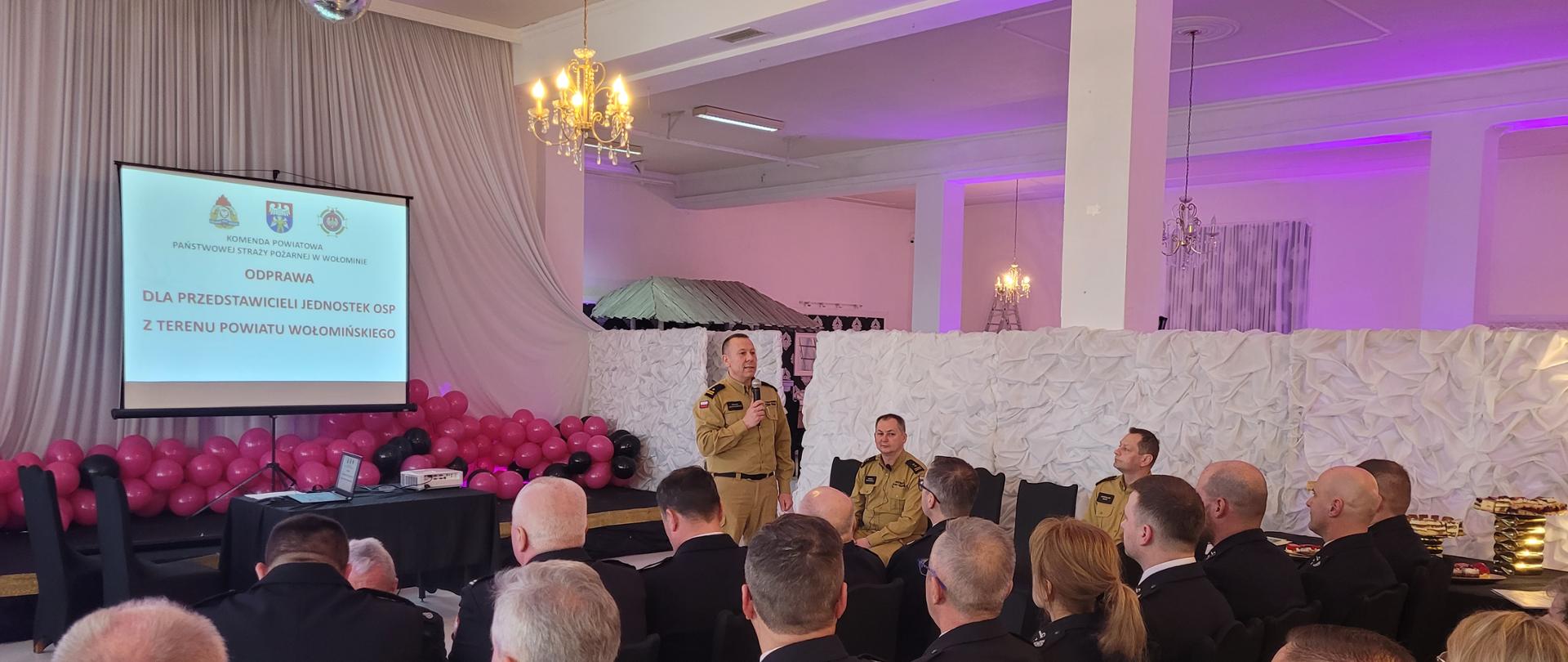 Zdjęcia przedstawiają strażaków OSP oraz PSP podczas narady rocznej w Sali bankietowej. Strażacy siedzą na krzesłach w mundurach wyjściowych. Funkcjonariusze PSP przedstawiają tematy omawiane podczas narady.