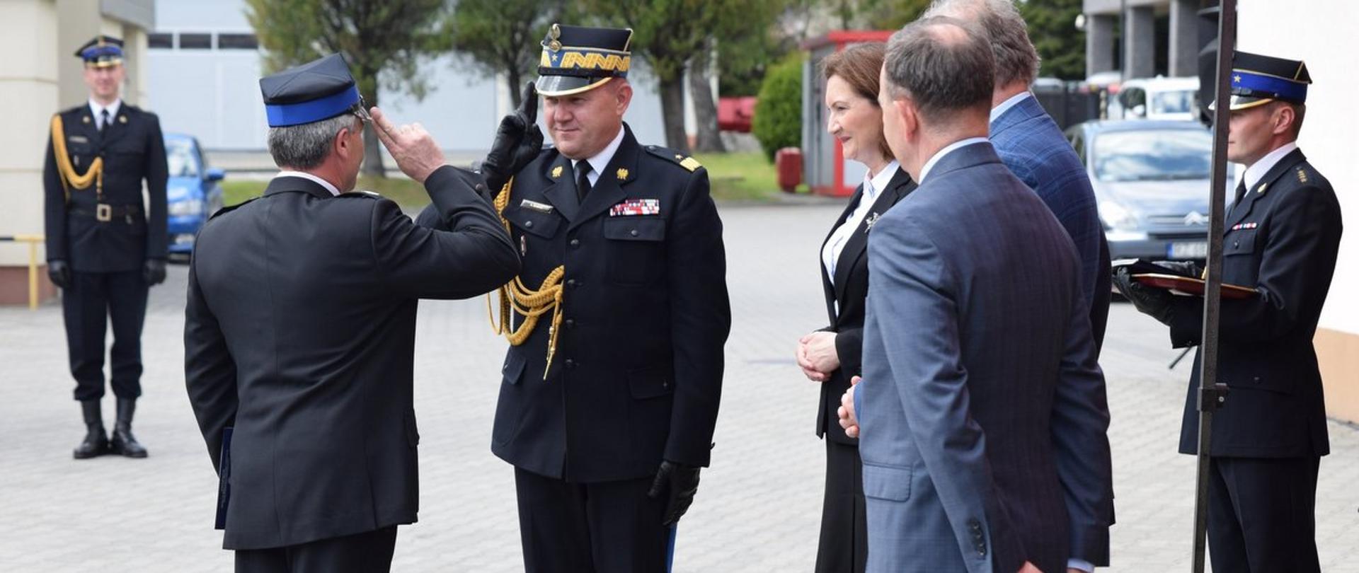 Na zdjęciu strażak w mundurze generała PSP oddaje salut strażakowi ochotnikowi. Obydwaj ubrani są w mundury galowe. Obok stoją za-proszeni goście. 