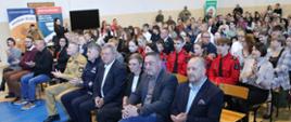 Szerokie ujęcie widowni zgromadzonej w hali sportowej podczas spotkania profilaktyczno-edukacyjnego – w pierwszym rzędzie siedzą oficjele cywilni, przedstawiciele Wojewody Świętokrzyskiego oraz funkcjonariusze Policji i Państwowej Straży Pożarnej.