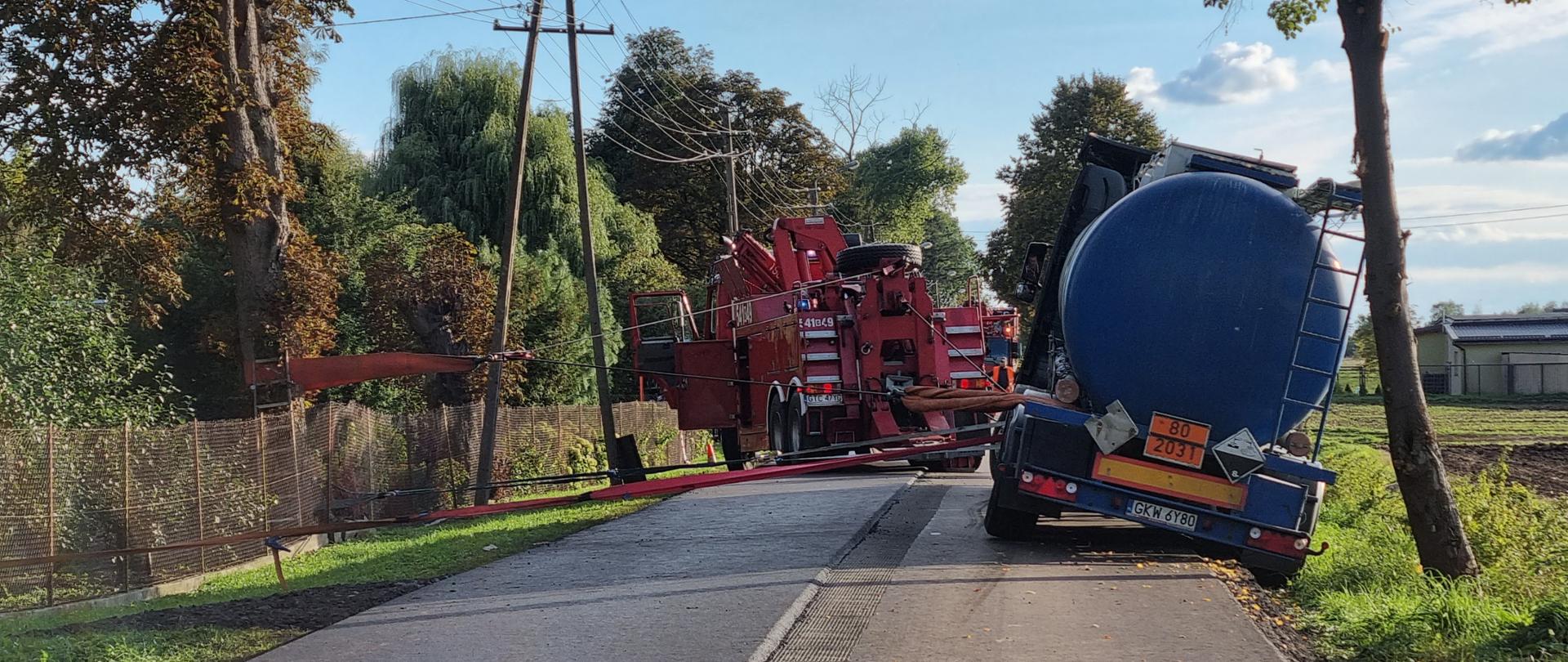 Przechylona cysterna stoi jedna strona na poboczu a drugą na jezdni, zabezpieczona pasami, trwa przygotowanie do jej wyciagnięcia
