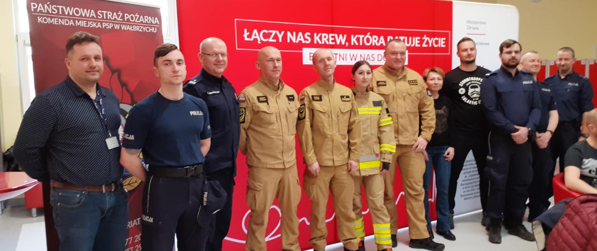 Policjanci w granatowych mundurach oraz strażacy w mundurach koloru piaskowego pozują do zdjęcia na tle czerwonego baneru