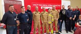 Policjanci w granatowych mundurach oraz strażacy w mundurach koloru piaskowego pozują do zdjęcia na tle czerwonego baneru