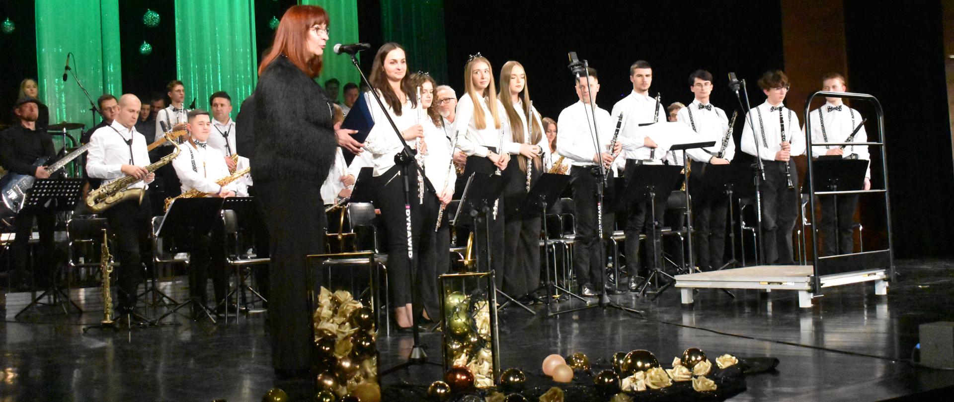 Na zdjeciu widać Dyrektor PSM Ist. W Jaśle stojącą przy mikrofonie na scenie Jasielskiego Domu Kultury w Jaśle podczas Koncertu Noworocznego. Obok niej ustawieni są młodzi muzycy orkiestry dętej, trzymający instrumenty i stojący przy pulpitach z nutami. W tle widoczna jest pełna obsada orkiestry oraz zielona, dekoracyjna scenografia, a przed sceną znajdują się eleganckie noworoczne ozdoby.