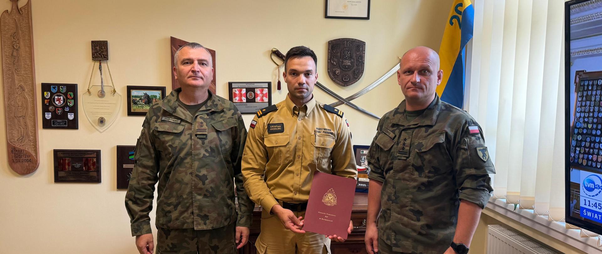 Na zdjęciu widać trzech mężczyzn stojących w biurze. Dwóch z nich, po lewej i prawej stronie, ubranych jest w mundury wojskowe w kamuflażu, a mężczyzna w środku ma mundur strażacki w kolorze piaskowym i trzyma w dłoniach bordową teczkę z godłem straży pożarnej. W tle widoczne są ściany ozdobione odznakami, herbami, fotografiami i mieczami, a także fragment flagi żółto-niebieskiej z herbem. Po prawej stronie kadru znajduje się telewizor wyświetlający fragment programu informacyjnego.