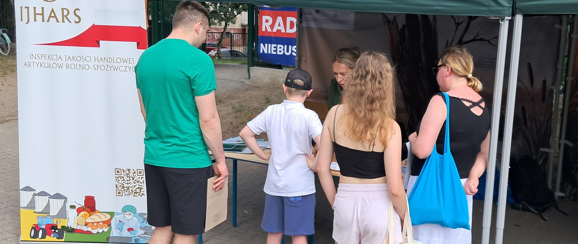 Stoisko IJHARS podczas festynu. Inspektorka rozmawia z trzema osobami dorosłymi i jednym dzieckiem, wyjaśniając zasady znakowania żywności oraz znaczenie informacji umieszczanych na etykietach produktów.