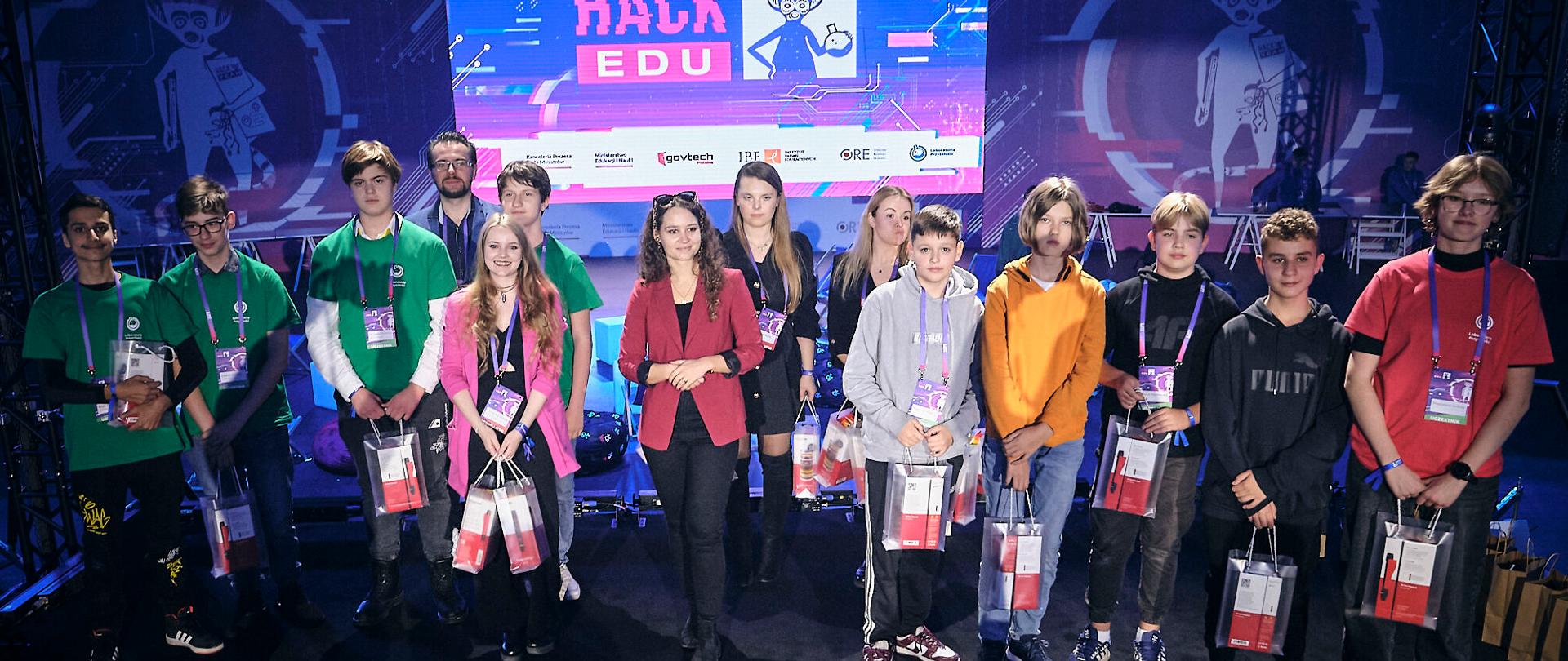 Grupa kolorowo ubranej młodzieży stoi na czarnej podłodze, w ciemnym pomieszczeniu, za nimi duży świecący na niebiesko i fioletowo monitor z napisem HackEdu.