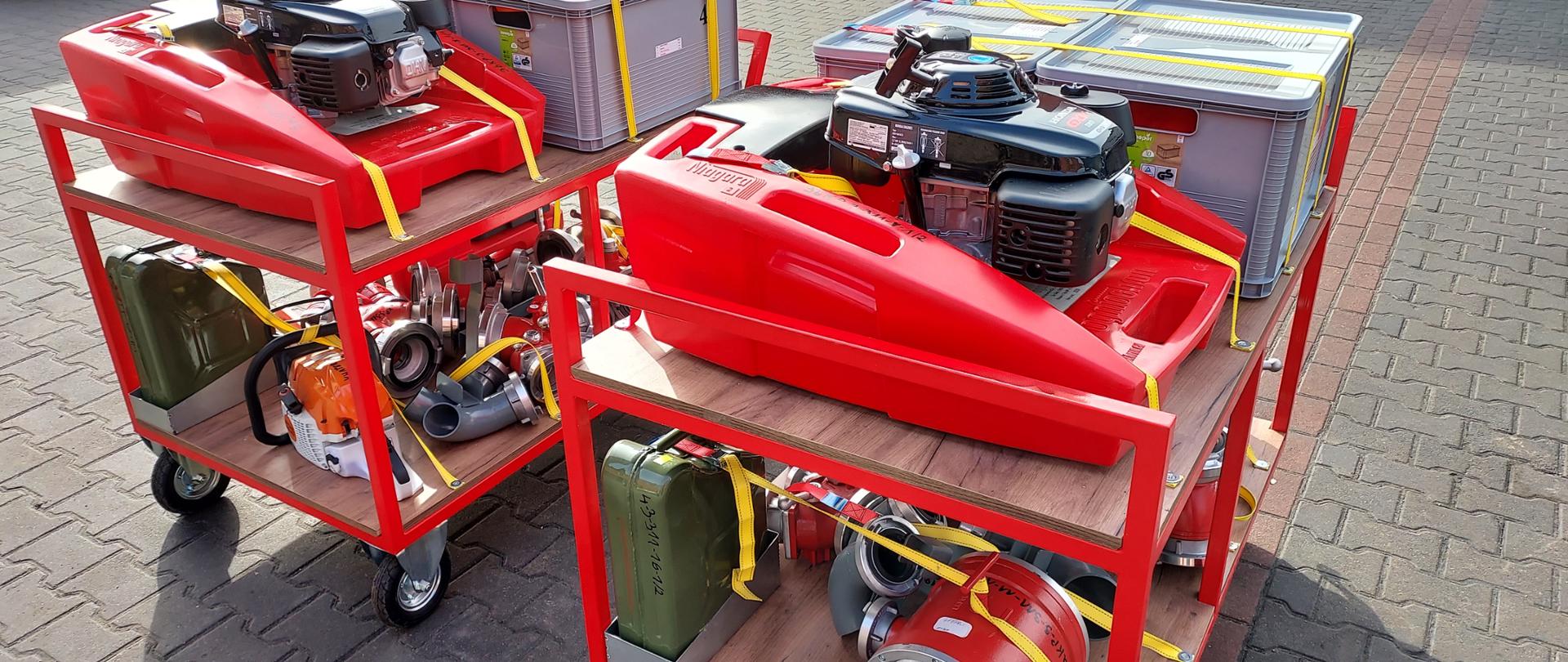 Słonecznie.Plac strażnicy. Na kostce brukowej stoją dwa kompletne wózki ze sprzętem pożarniczym m.in. czerwone motopompy pływające, szare skrzynki, zielone kanistry z paliwem, czerwona armatura pożarnicza, biało-pomarańczowa pilarka do drewna.