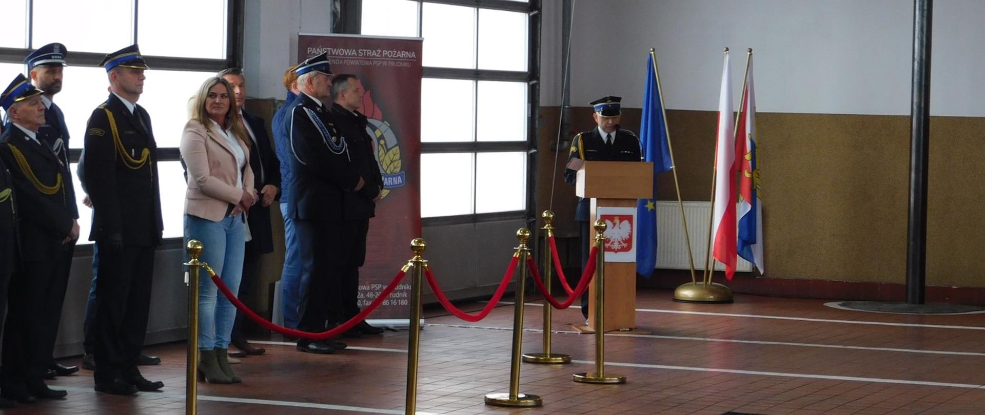Zdjęcie przedstawia strażaka podczas przemowy przy mównicy na uroczystym apelu z okazji Dnia Strażaka.
Na zdjęciu widoczni z lewej strony zaproszeni goście.