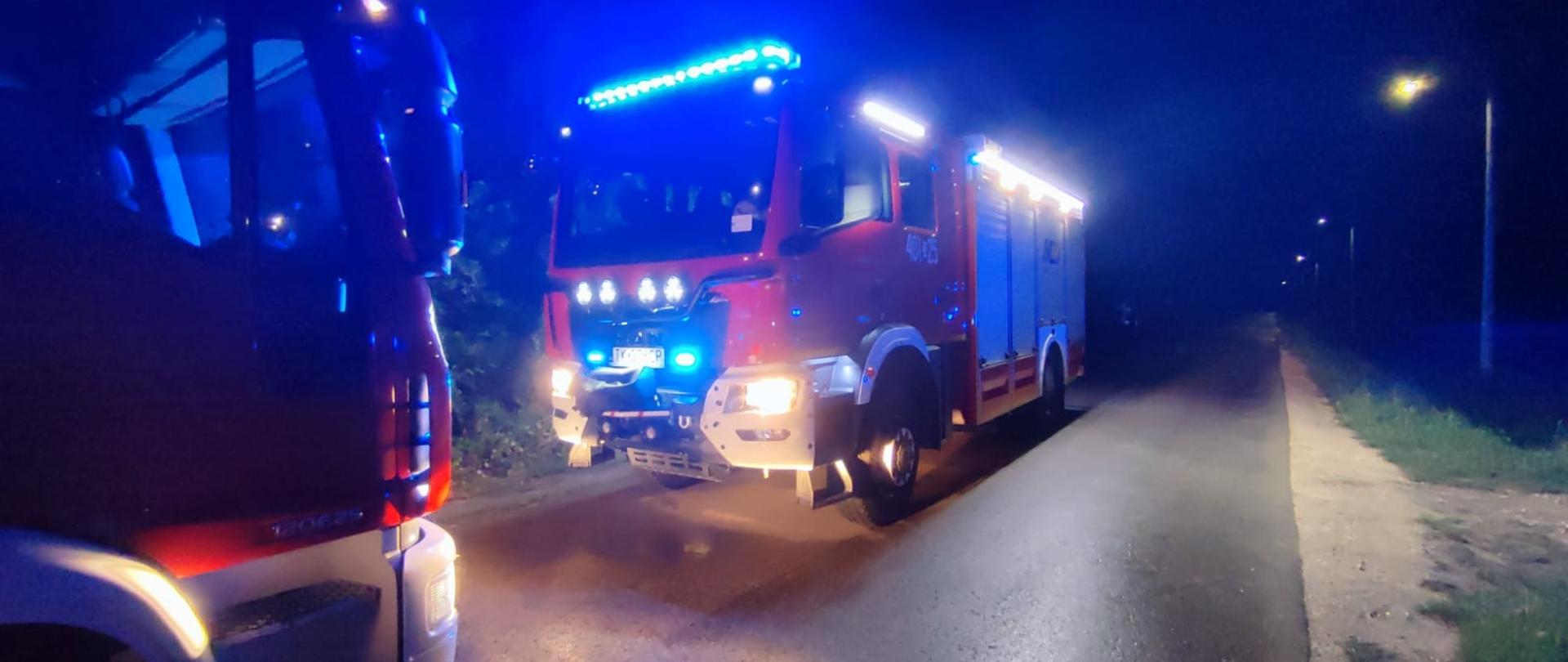 Zdjęcie ukazuje wozy strażackie zaparkowane na ciemnej drodze lub poboczu w nocy. Pojazdy mają włączone niebieskie światła ostrzegawcze (syngnalizację świetlną) oraz reflektory, co wskazuje na trwającą akcję ratowniczo-gaśniczą w okolicy.