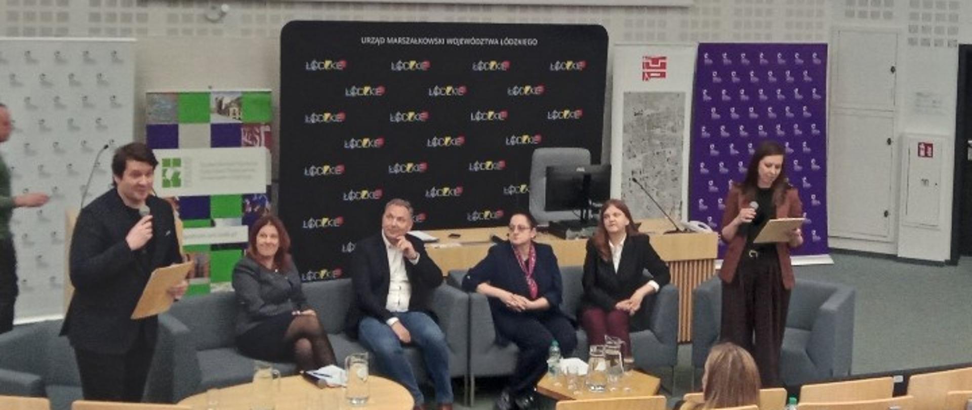 Na zdjęciu widać salę wykładową z ustawioną z przodu sceną. Kilka osób siedzi na kanapach i fotelach, uczestnicząc w panelu dyskusyjnym. Po lewej stronie stoi mężczyzna z mikrofonem i kartką w ręku, prawdopodobnie prowadzący spotkanie. Po prawej stronie stoi kobieta, również z mikrofonem i notatkami. Za panelistami znajduje się ekran projekcyjny z wyświetloną prezentacją oraz banery z logo „Łódzkie”. 