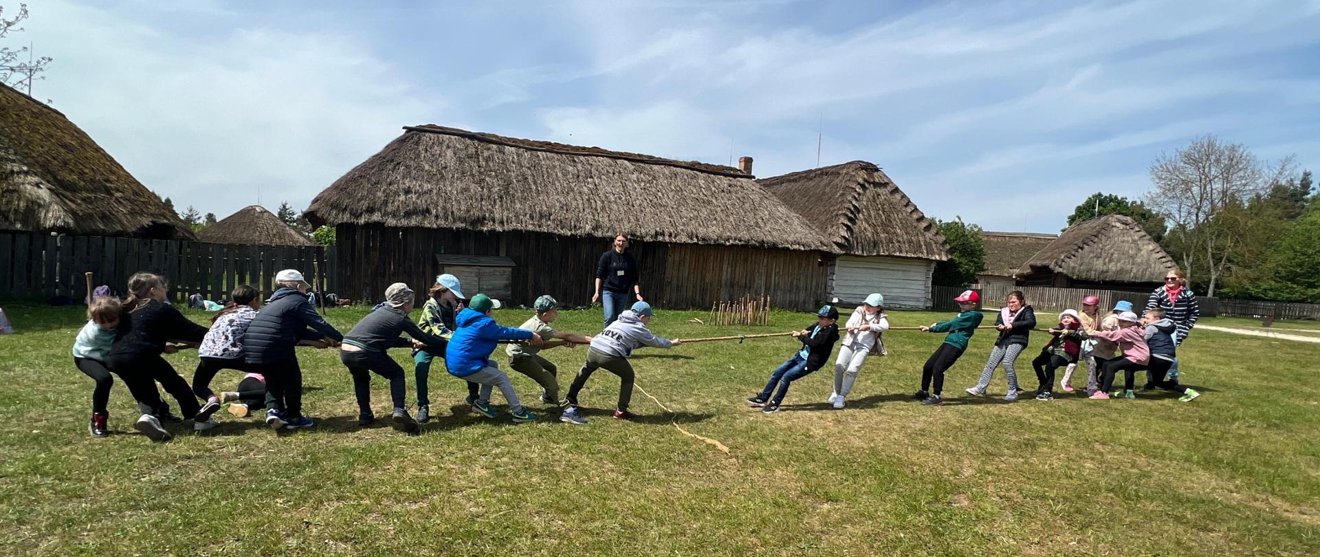 Na polanie dzieci ustawione w dwóch drużynach, naprzeciwko siebie, bawią się w przeciąganie liny. W tle znajdują się stare chałupy, pokryte strzechą, dalej po prawej stronie drzewa, niebo ma piękny, błękitny kolor.
