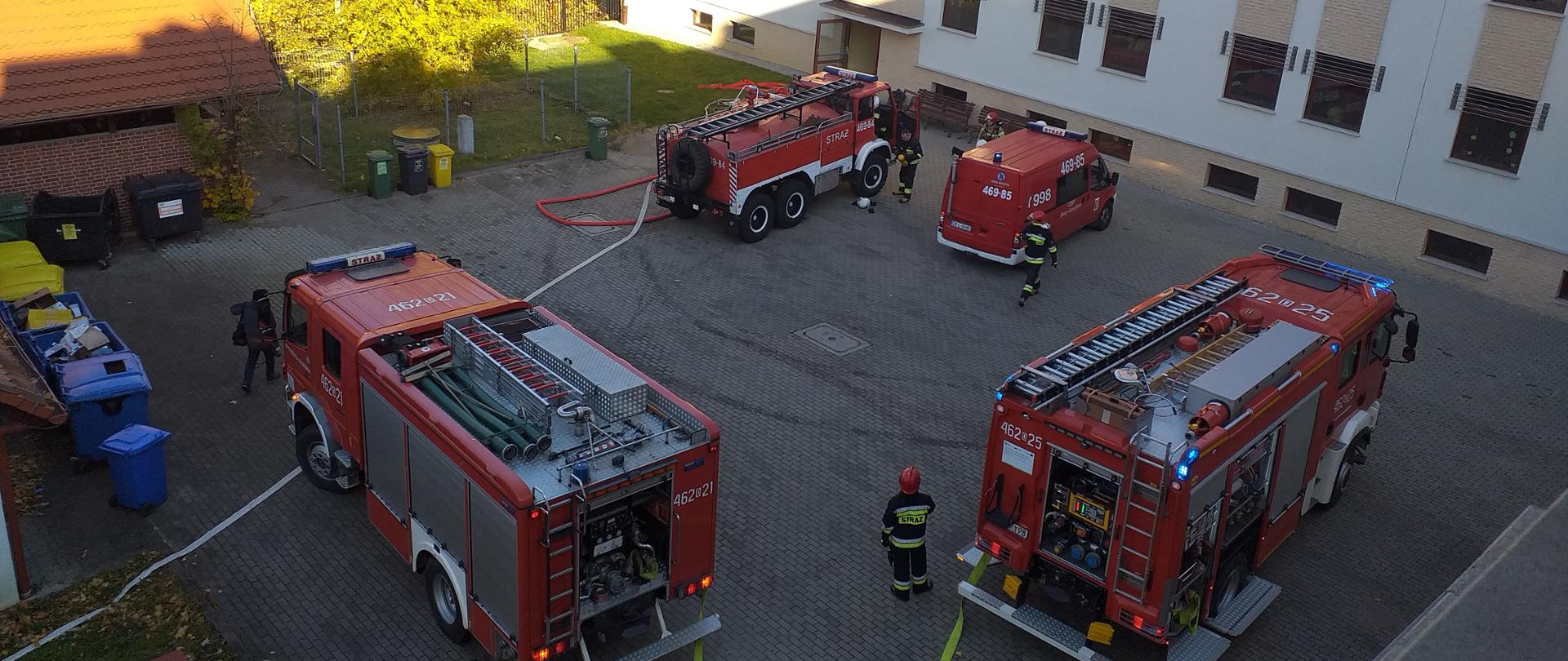 Widok na plac przy Wojewódzkim Centrum Psychiatrii Długoterminowej w Stroniu Śląskim. Na placu stoją cztery samochody pożarnicze oraz strażacy przygotowywujący się do ćwiczeń.