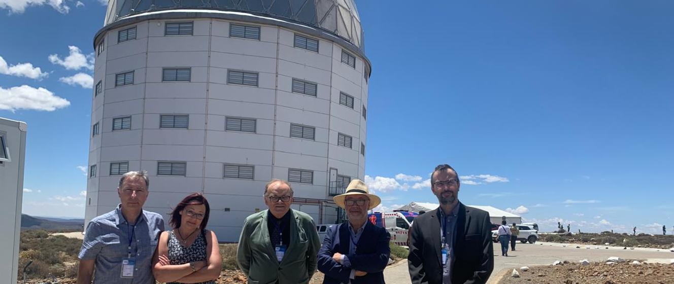Grupa pięciu osób (jedna kobieta i czterech mężczyzn) stoi przed budynkiem Obserwatorium Astronomicznego (SAAO) w Sutherland