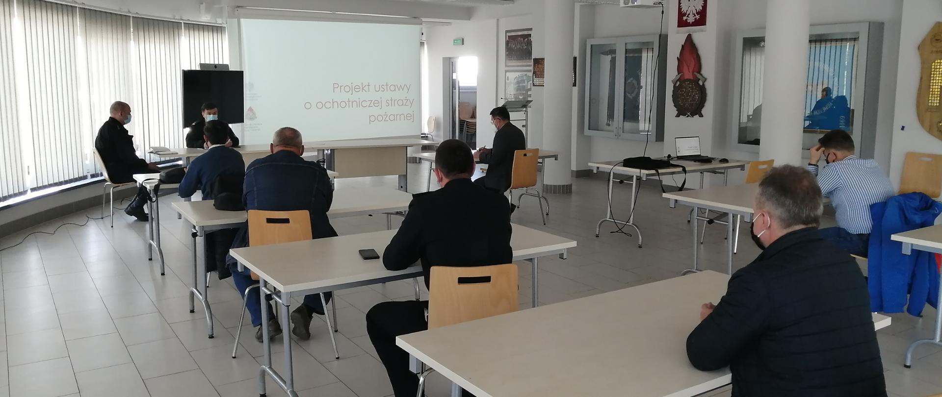 Dwóch mężczyzn ubranych w czarny mundur strażacki oraz sześciu mężczyzn ubranych w ubrania cywilne tj,: koszule oraz kurtki, na twarzy mają ubraną maseczkę chirurgiczną koloru biało niebieskiego oraz czarne, siedzą przy stołach na Sali konferencyjnej. Mężczyźni siedzą przy stolikach w odległości każdy od siebie ok. 2 metry. W tle na ekranie wyświetlana jest prezentacja dot. ustawy o OSP.