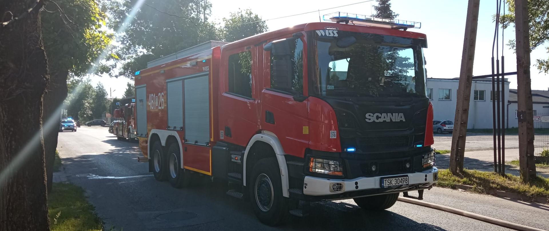 Zdjęcie przedstawia ciężki samochód gaśniczy Scania będący przed wjazdem na teren zakładu stolarni. Za nim stoi drabina mechaniczna i radiowóz policyjny. Wszystkie pojazdy mają włączone sygnały świetlne.