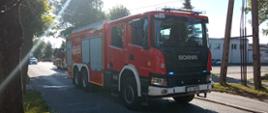 Zdjęcie przedstawia ciężki samochód gaśniczy Scania będący przed wjazdem na teren zakładu stolarni. Za nim stoi drabina mechaniczna i radiowóz policyjny. Wszystkie pojazdy mają włączone sygnały świetlne.
